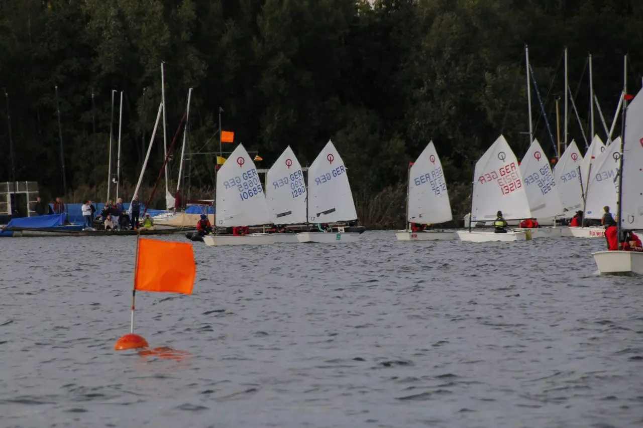 Optimisten-Jollen mit weißen Segeln hinter einer orangefarbenen Markierung